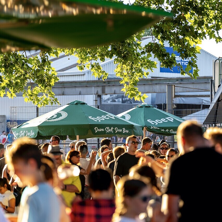 Besucherinnen und Besucher beim HafenKulturFest 2025 am Karlsruher Rheinhafen, mit Biergarten-Atmosphäre, Sonnenschirmen und sommerlicher Stimmung im Hafenumfeld.