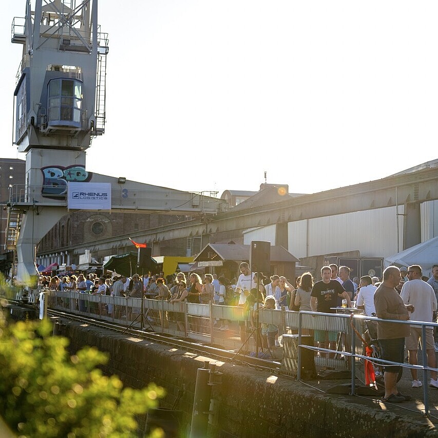 Besucherinnen und Besucher beim HafenKulturFest 2025 am Karlsruher Rheinhafen entlang der Kaimauer, mit Bühnenbereich, Industriekran und sommerlicher Festivalatmosphäre.